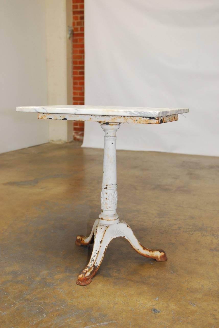 Antique French white marble-top bistro table featuring an iron pedestal base with tripod legs. Made in the Art Nouveau taste with floral swag detail on the column and a distressed white finish. Topped by a square piece of white Carrara marble with