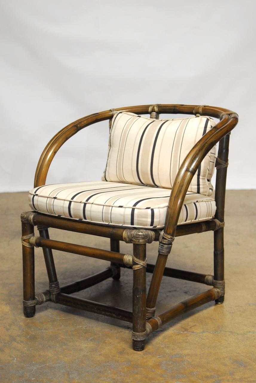 Mid-Century pair of barrel back armchairs constructed from bamboo rattan featuring an open fretwork backrest and reinforced with rawhide strapping. Graceful arms curve around the entire frame and attach at the stretchers with a cushion seat and down