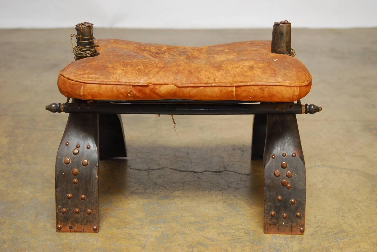 Beautifully distressed Moroccan camel saddle stool featuring a padded leather cushion. The frame is reinforced with rawhide strapping and embellished with decorative metal studs. The tops of the wooden frames end with round handles. Great authentic