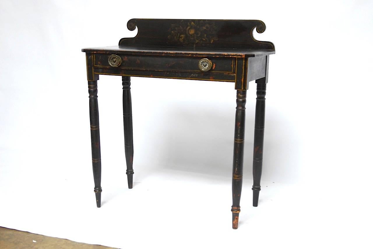 19th century Federal work table or desk. Made in the style of Lambert Hitchcock. Featuring an ebonized finish decorated with delicate yellow accent stripes and a floral motif painted rear gallery. Fronted by a large drawer with two round brass pulls