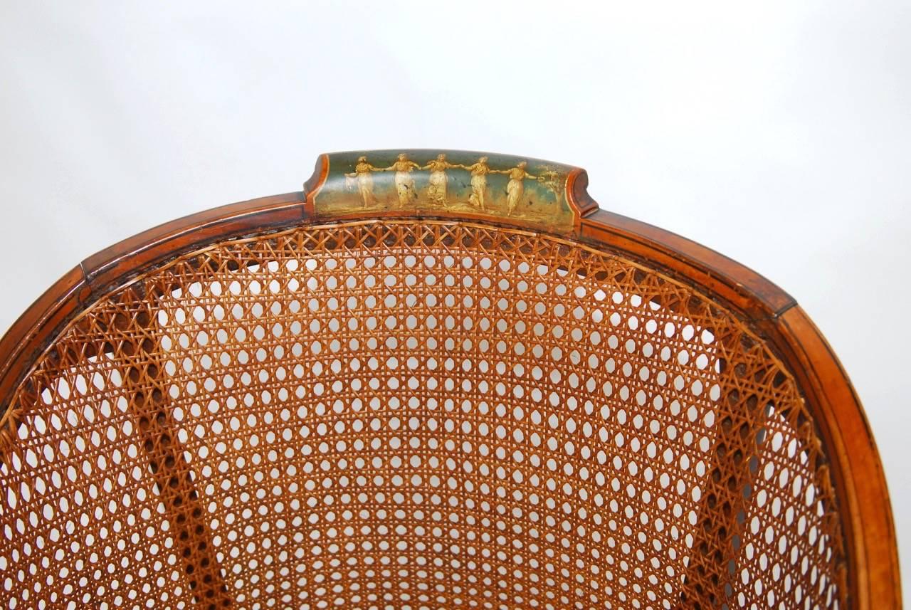 Unique English paint decorated and caned armchair made in the Edwardian period featuring neoclassical style decoration on the crest and seat. Made with a barrel back shape and having caned sides and seat supported by tulip legs and feet. Thick