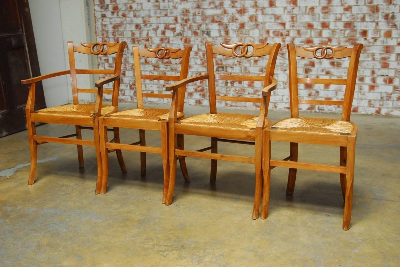 Attractive set of four French country dining chairs with rush seats featuring a hand-carved fruitwood frame. Set consists of two armchairs and two side chairs all ladder back style with a decorative carved double winged circle crest rail. The seats