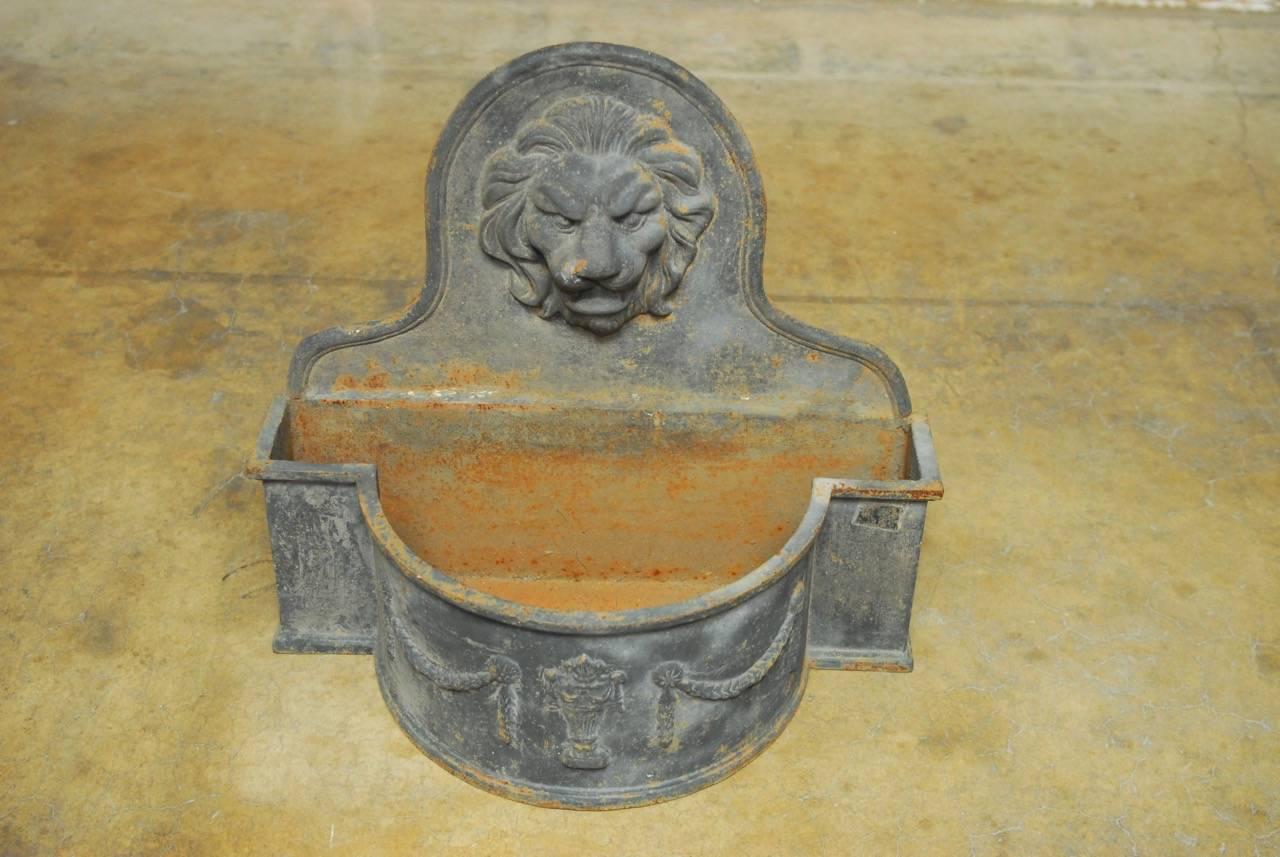 Patinated Louis XVI style cast iron wall fountain featuring a lion's head motif and swag neoclassical decoration. The fountain has a bowed demilune form in the front, and a gently curved crest. The cast iron has a rich, patinated finish.