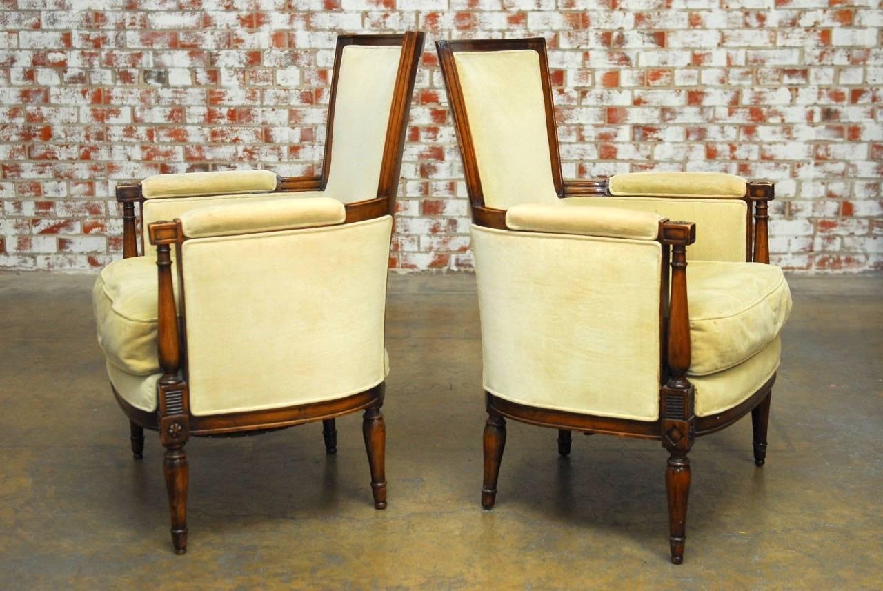 Grand pair of French Louis XVI walnut bergere armchairs made in the manner of Maison Jansen. Featuring a padded square back frame with a barrel shape. The walnut frame has column supports on the front arms decorated with rosettes and a bowed front
