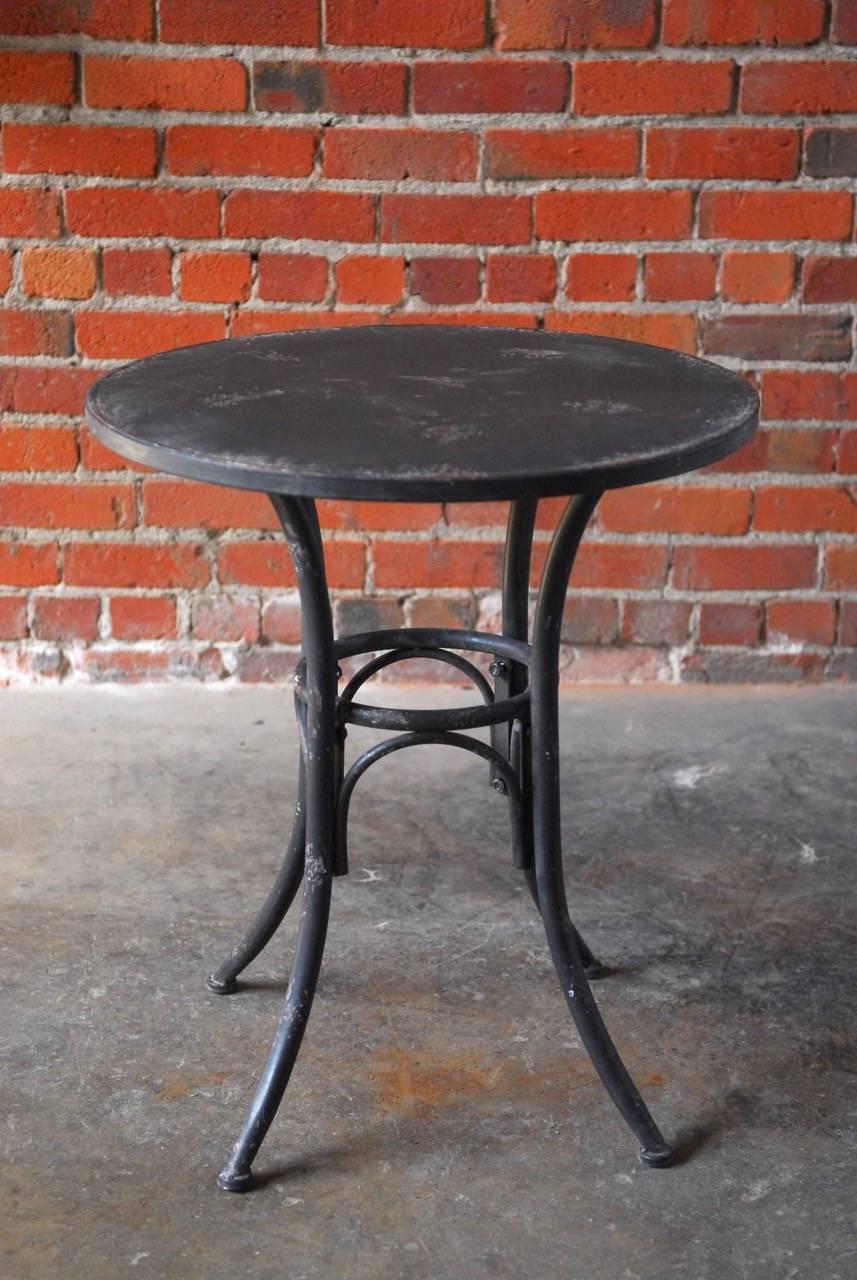 Metal patio bistro table with a painted and distressed black finish on the metal. Supported by four legs with a round stretcher.