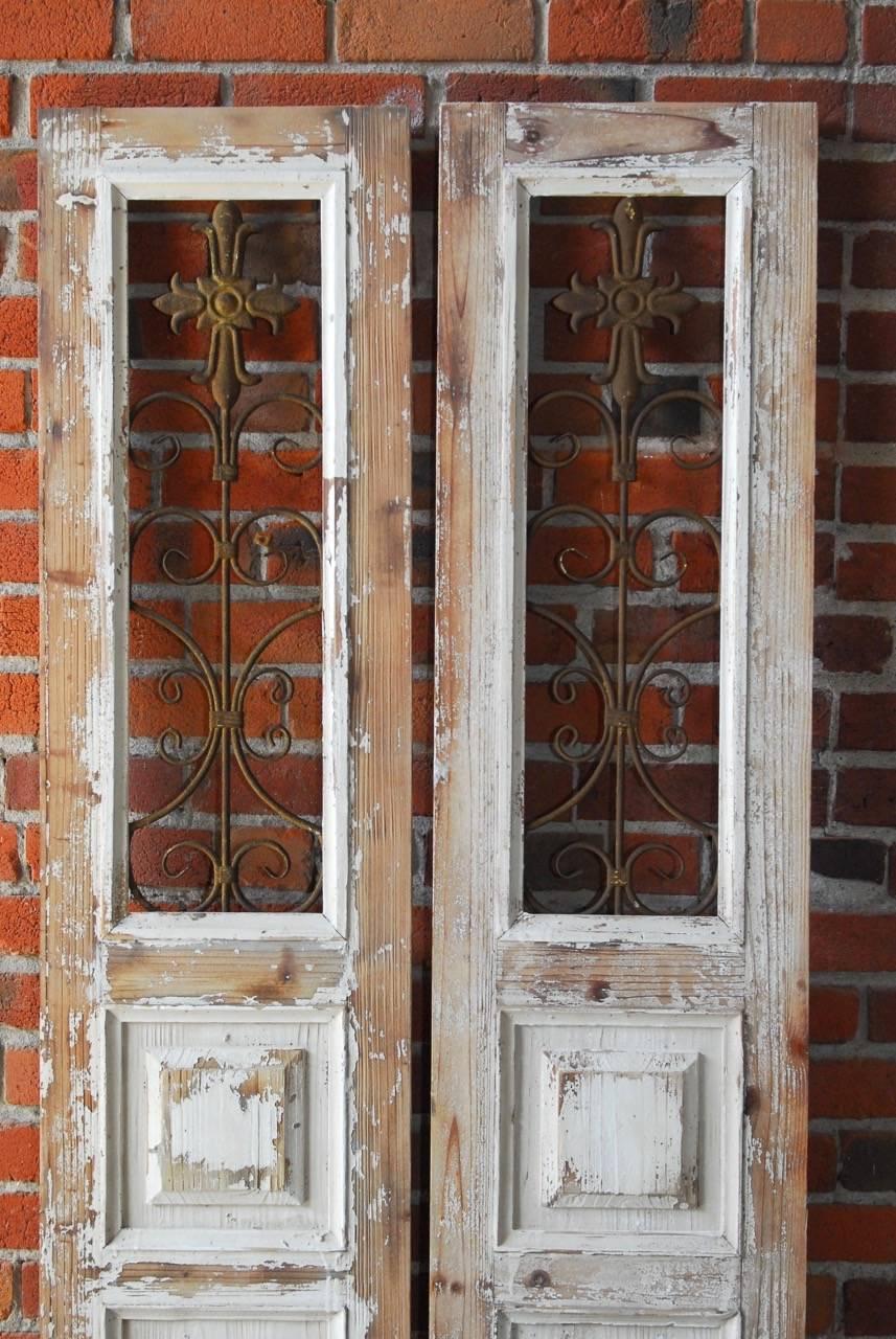 Distressed pair of pinewood window shutters or door pockets with scrolled ironwork grills. Doors have paneled bottoms and decorative metal work open window tops with a cross motif and a patinated metal finish. Remnants of white paint on wooden