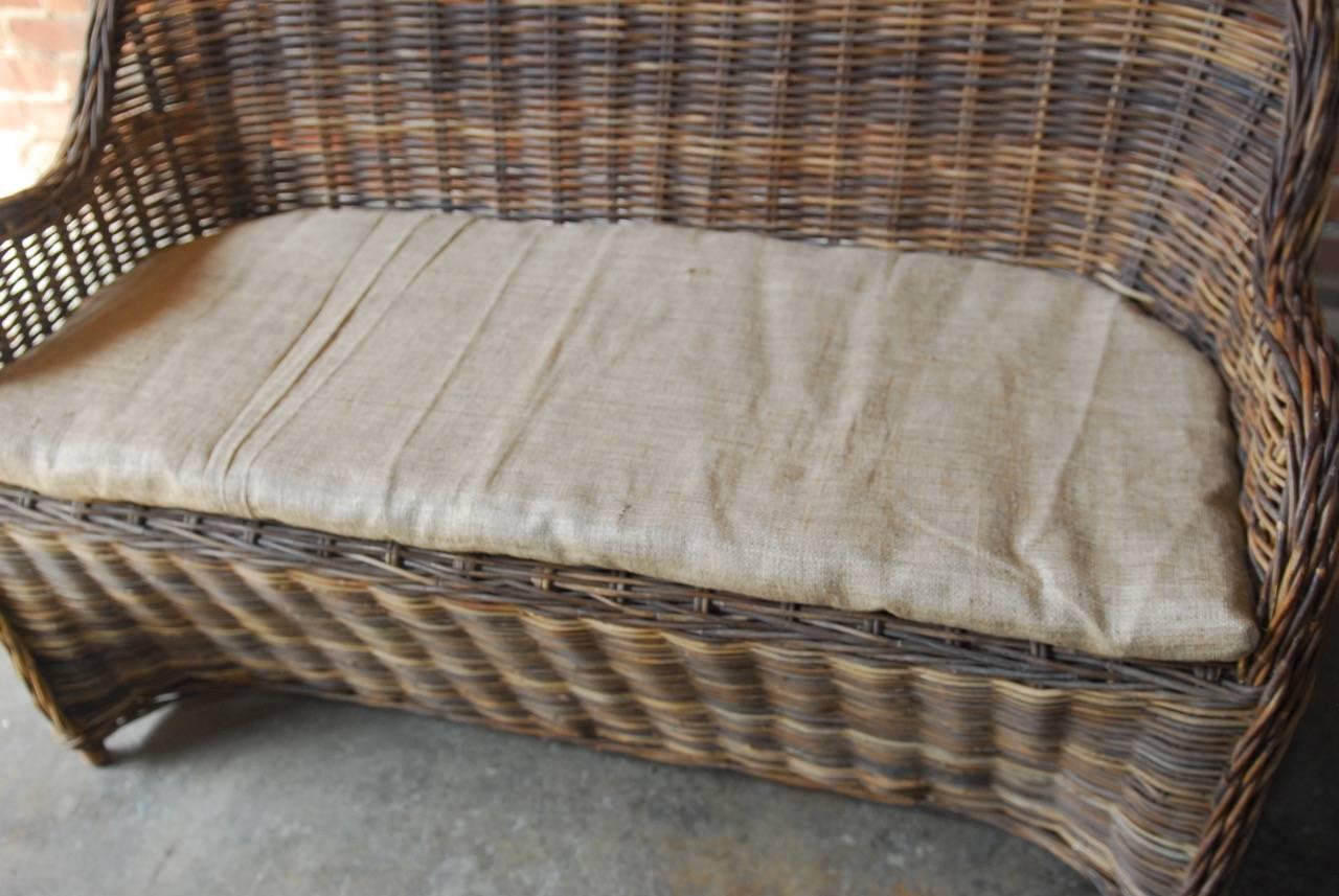 Distinctive organic modern woven rattan and wicker settee or dining bench seating. Constructed from a natural rattan frame with graceful braided arms and back. Topped with a thin burlap seating cushion. Makes an excellent bench style seating for a