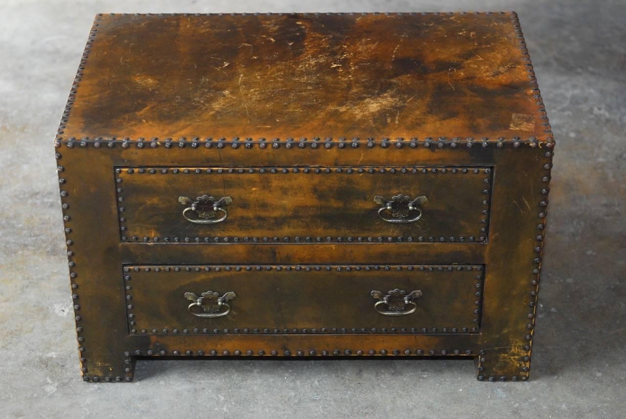 Diminutive tabletop chest of drawers featuring a handsome leather cladding bordered with brass metal nail head trim. The leather has a distressed, rustic patina with scuffs and rich wear that showcases the lovely colors in the leather. Fronted by