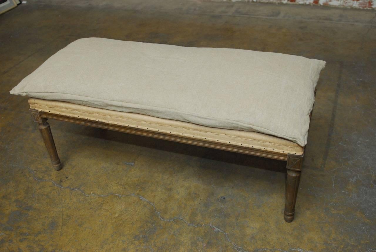 Distinctive French Louis XVI style oak bench featuring an exposed nailhead burlap upholstery with hand stitching. The bench has an organic linen pillow cushion on top and is supported by turned legs. Lovely farmhouse deconstructed style.