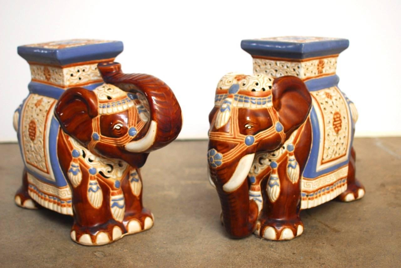 Playful pair of ceramic Asian elephant garden stools or drinks tables. Featuring caparisoned bodies with hand-painted saddles and costumes. One is depicted with its trunk up and the other is down. Vibrant colors with prosperity emblems on the seat