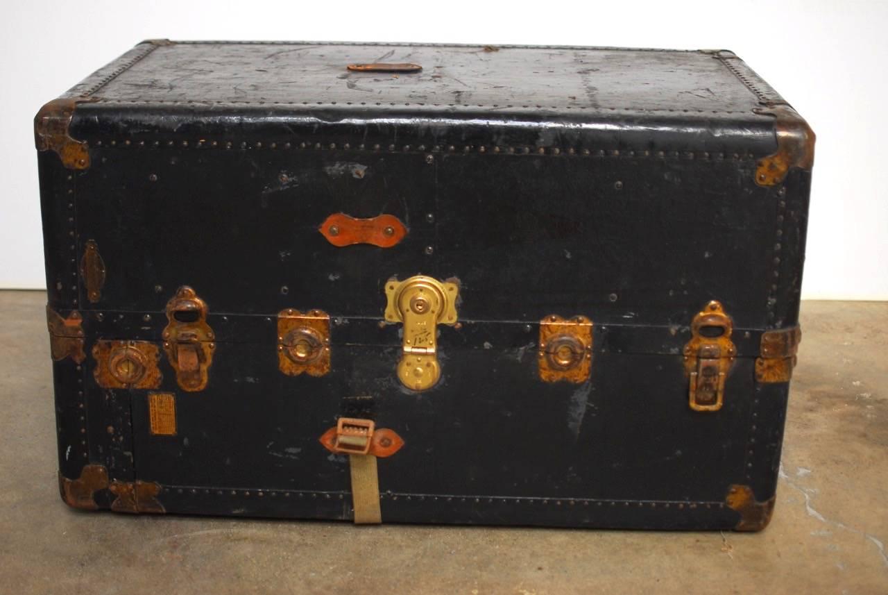 Black wardrobe steamer trunk made in the manner of Hartmann. Features a vulcanized fibre construction accented with brass studs and a canvas strap. Beautifully distressed and patinated finish with a leather handle on top. Makes a great coffee table.