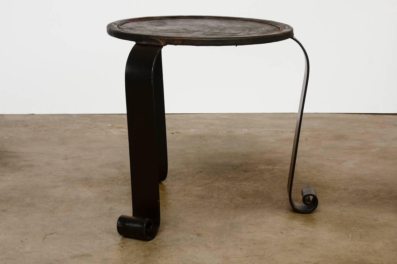 Rustic wrought iron leather top stool or drink table. Features a three leg design with decorative scrolled feet and a thick hand-stitched leather round top that is woven. Comfortable as a stool or strong enough to be used as a side table or drink