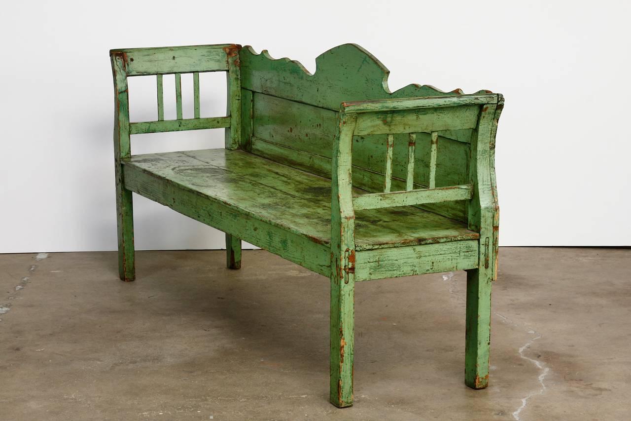 Rustic country French farmhouse pine bench or settle featuring a vintage green lacquered finish. Constructed with mortise and tenon joinery with a decorative scalloped back. The arms are scrolled and open with slats. The bench has a plank top seat