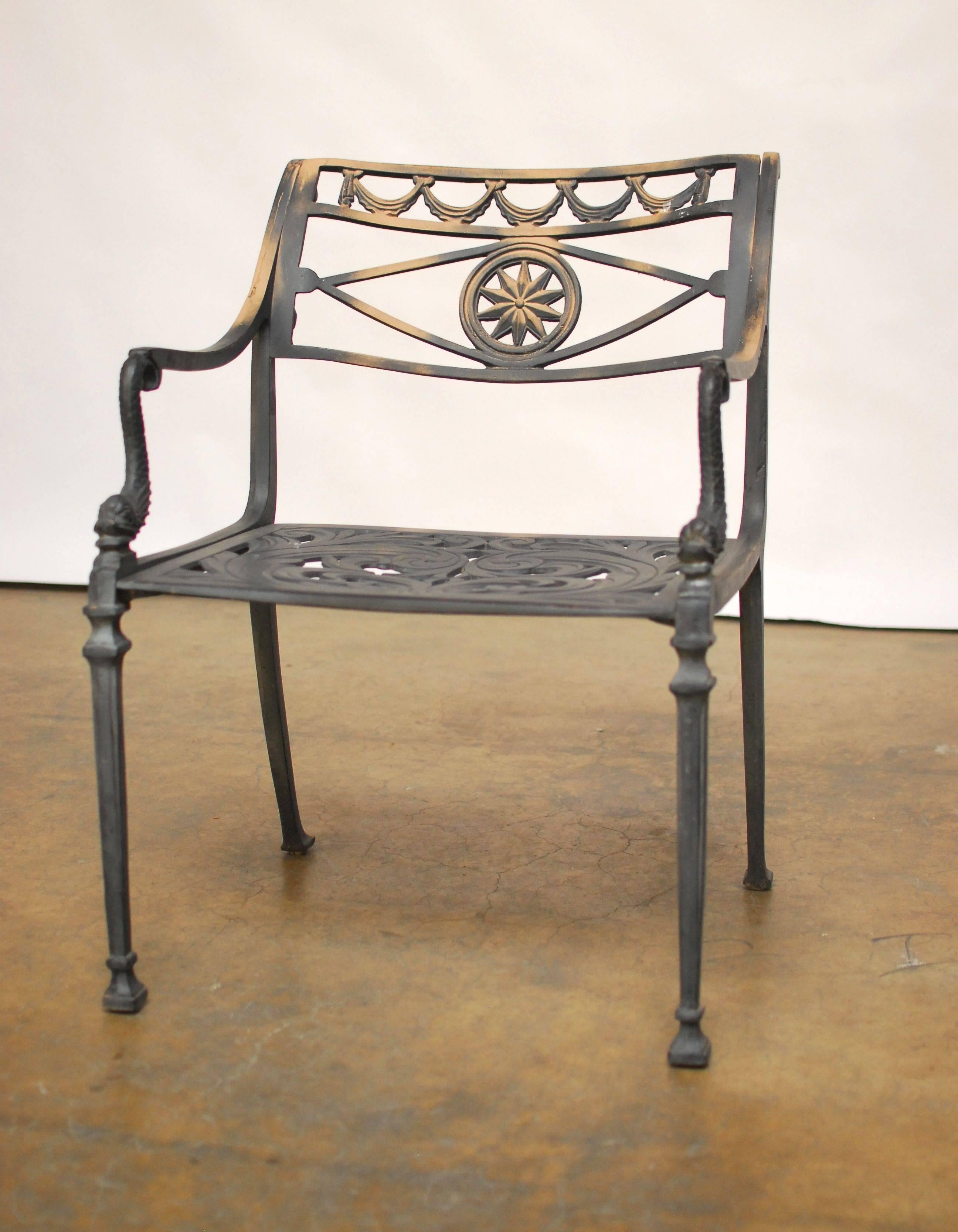 A set of four Directoire style garden armchairs made of cast metal featuring a sun medallion back and dolphin form arms. They have decorative pierced seats and detailed legs. They are a solid construction with a vintage matte patina finish. Very