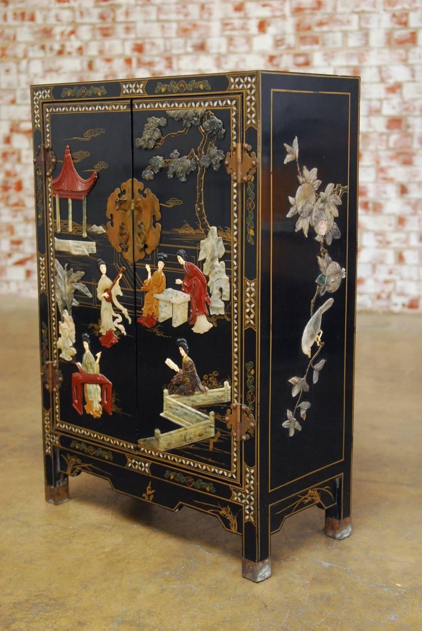 Stunning Chinese export black lacquer soapstone scholars cabinet fronted by two large doors above a shaped apron. The doors are decorated with intricately carved social scenes, pagodas, and trees. The sides are decorated with birds, fruit, and