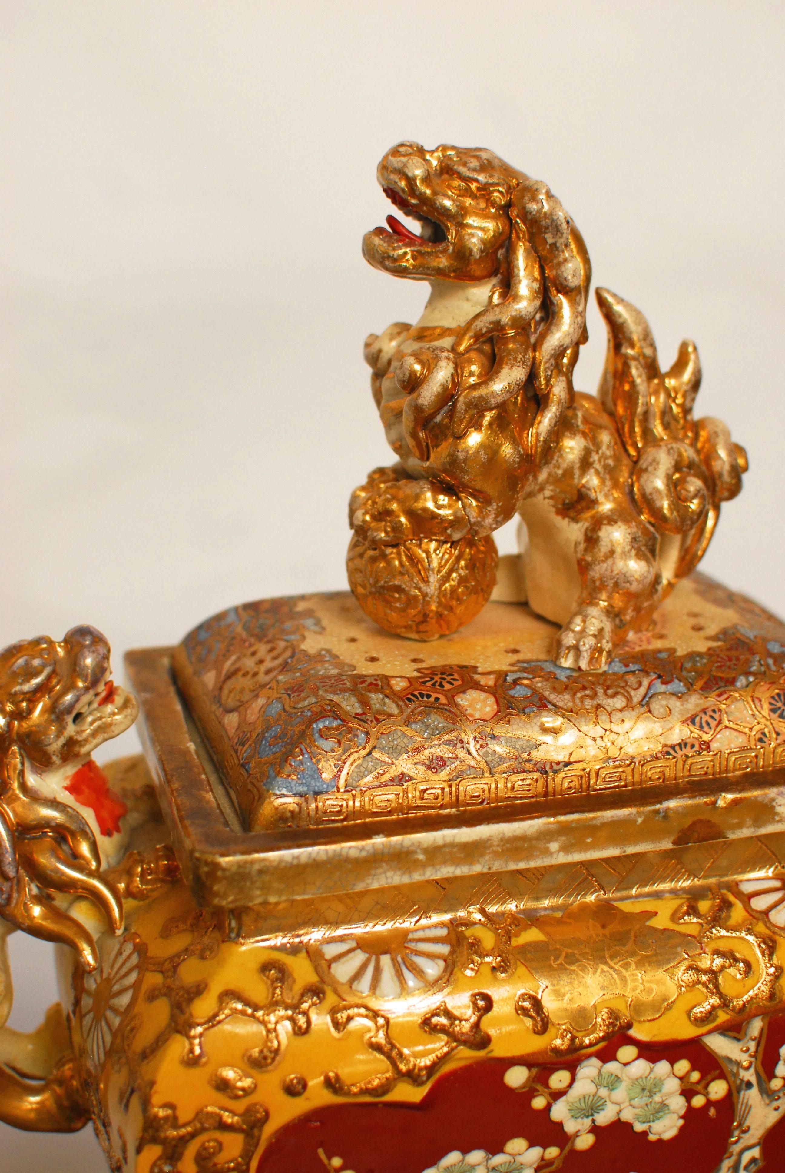 Large Japanese Satsuma ware censer featuring Shi-Shi handles and a rare square shaped bowl with four legs. The lid having a Foo-lion holding a ball with a Greek key pattern around the border. An abundance of gilt trim all-over. The brightly colored