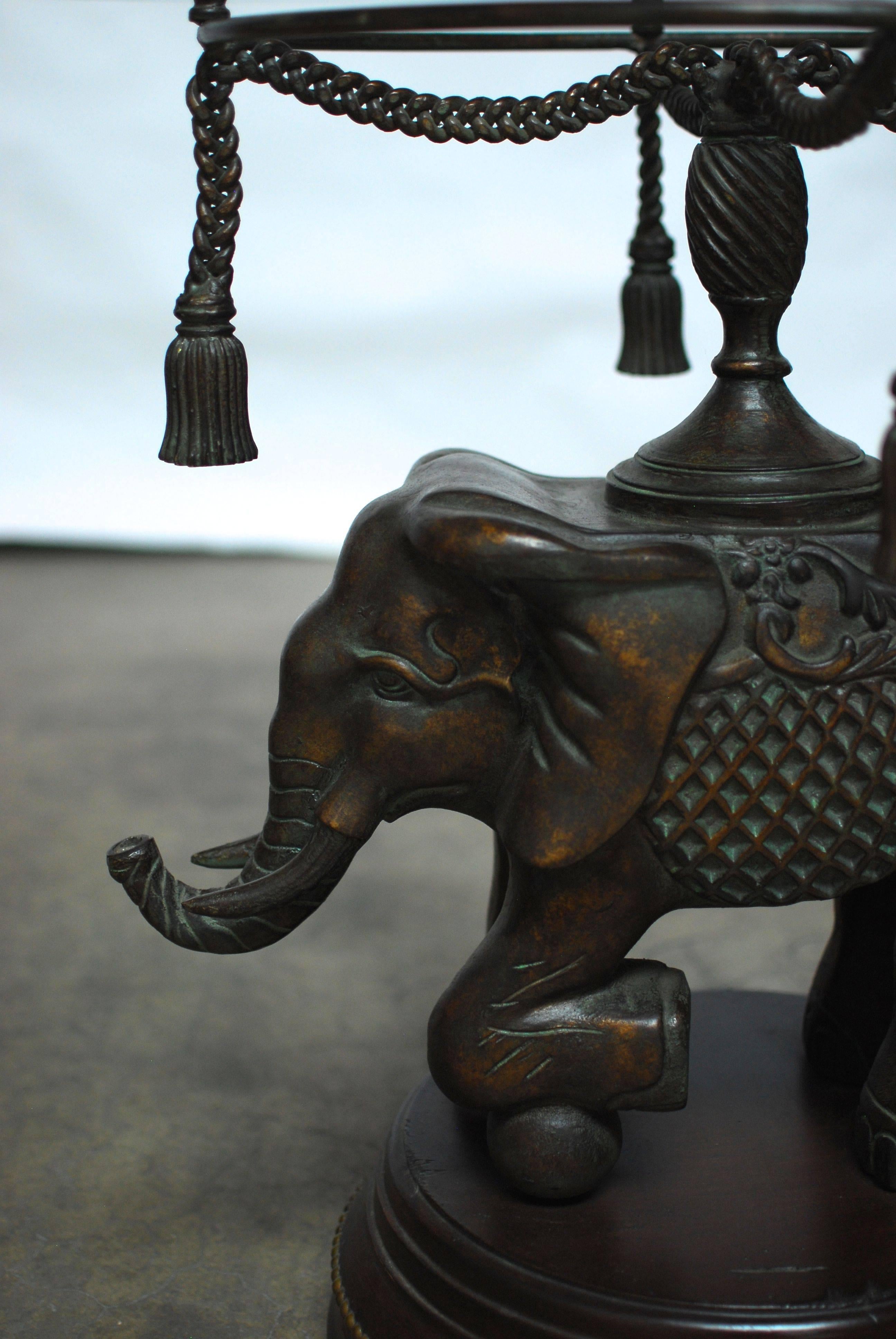 Bronze elephant standing on a carved wood plinth with decorative brass nailheads. Surmounted by a bronze pedestal halo with a swag faux rope terminating in four tassels. The trunk up elephant has tusks and a fretwork costume. This side table has a