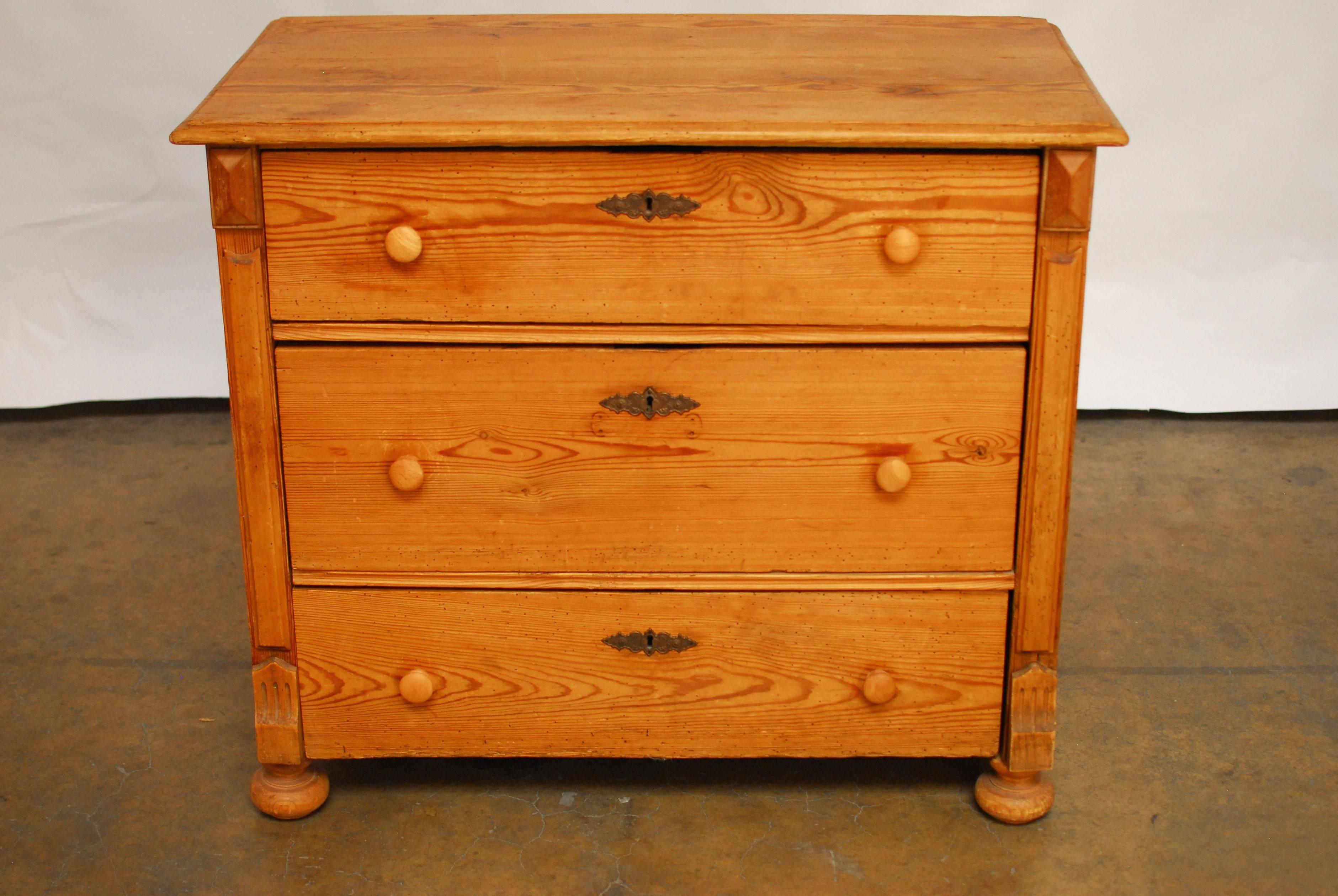 A rustic, 19th century French country pine three-drawer commode with a natural raw finish with a warm soft patina and worm holes on the vintage surfaces. Round wood pulls and decorative escutcheons add to the vintage look. This chest of drawers sits