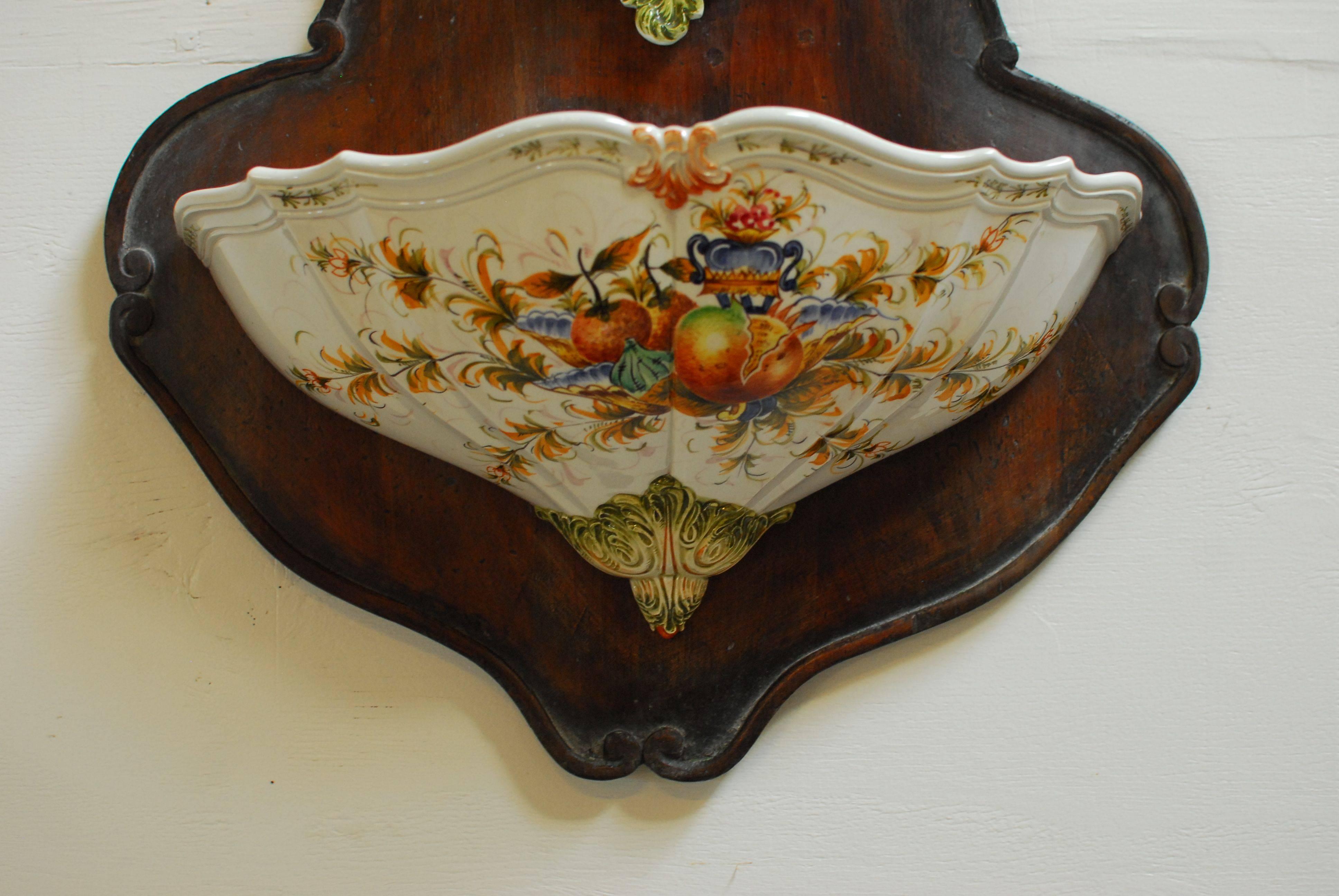 Rococo style Italian porcelain three piece lavabo hanging wall fountain mounted to a carved shaped base with a decorative edge. Top cistern has brass metal spigot and both pieces feature vibrant hand painted scenes with fruits, flowers, and bright