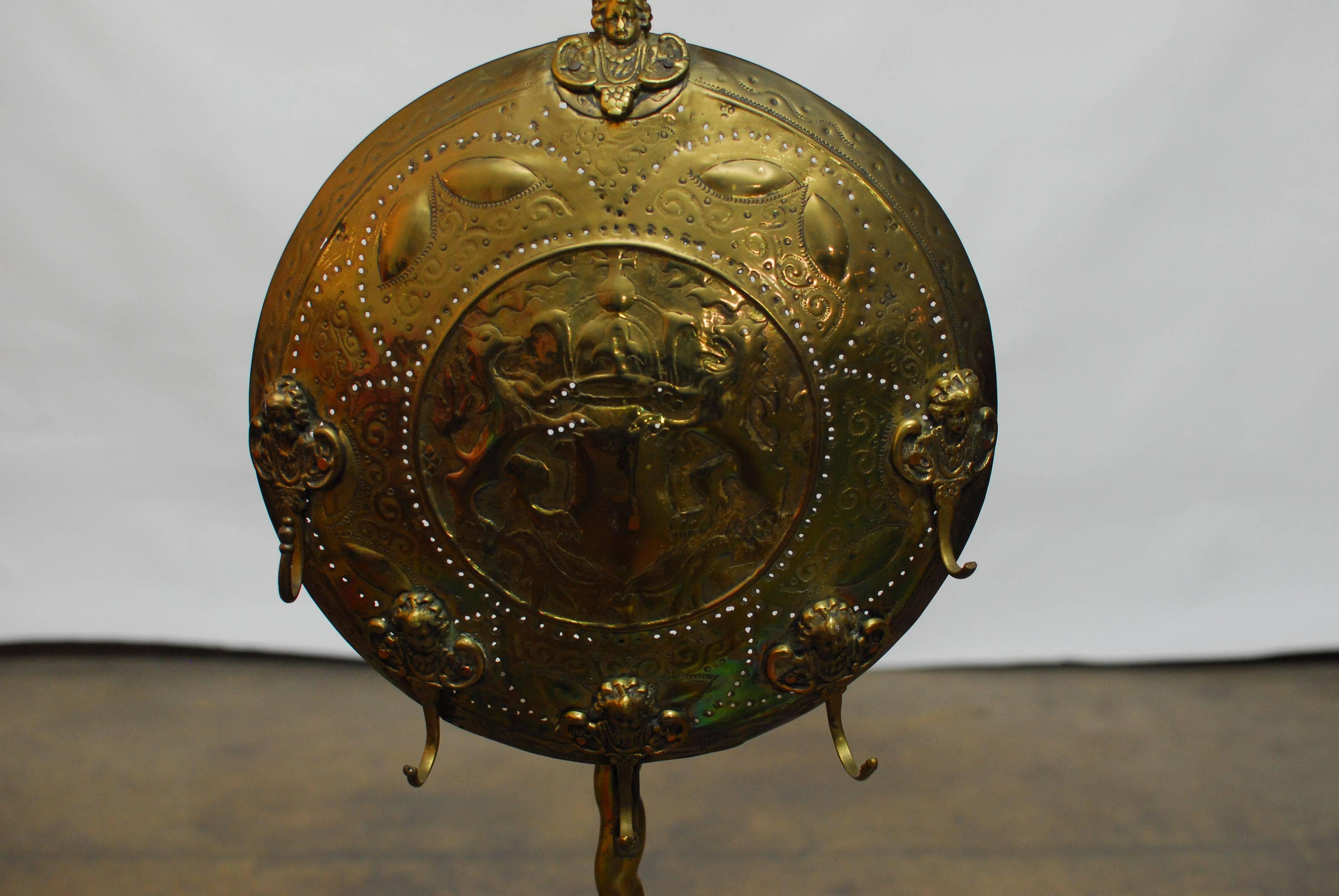 A rare brass fireplace tool stand with a candle holder featuring an intricate repoussé coat of arms with five cherubs each having a fire tool hook. The solid main shaft has a twist detail and sits on a tripod base with matching legs. Surmounted by a