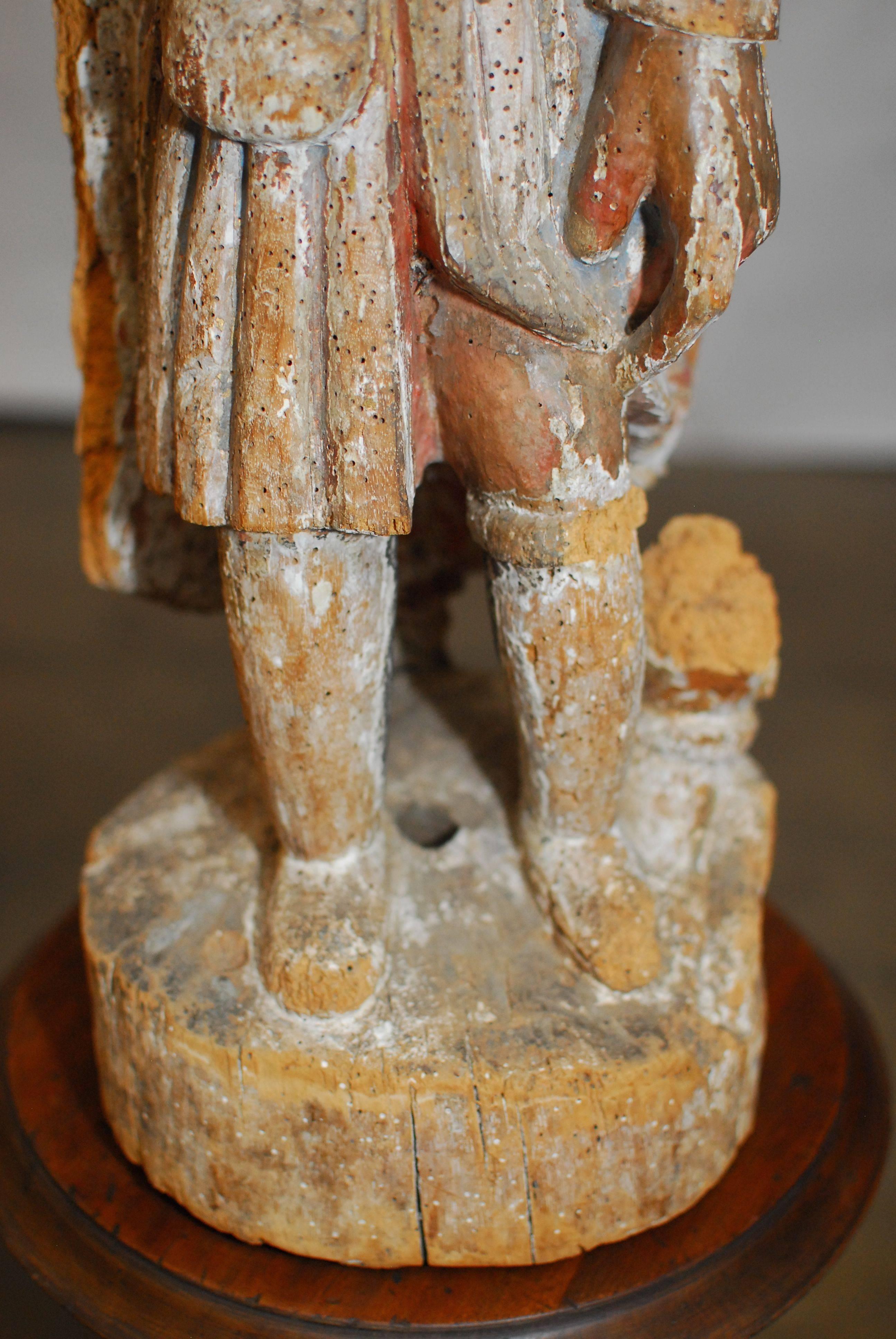 18th century polychromed wood folk art carving of a man depicted wearing a top hat and coat with some original color still visible. Minor losses and insect damage with age appropriate patina. Stands on a round wooden base. His right hand was