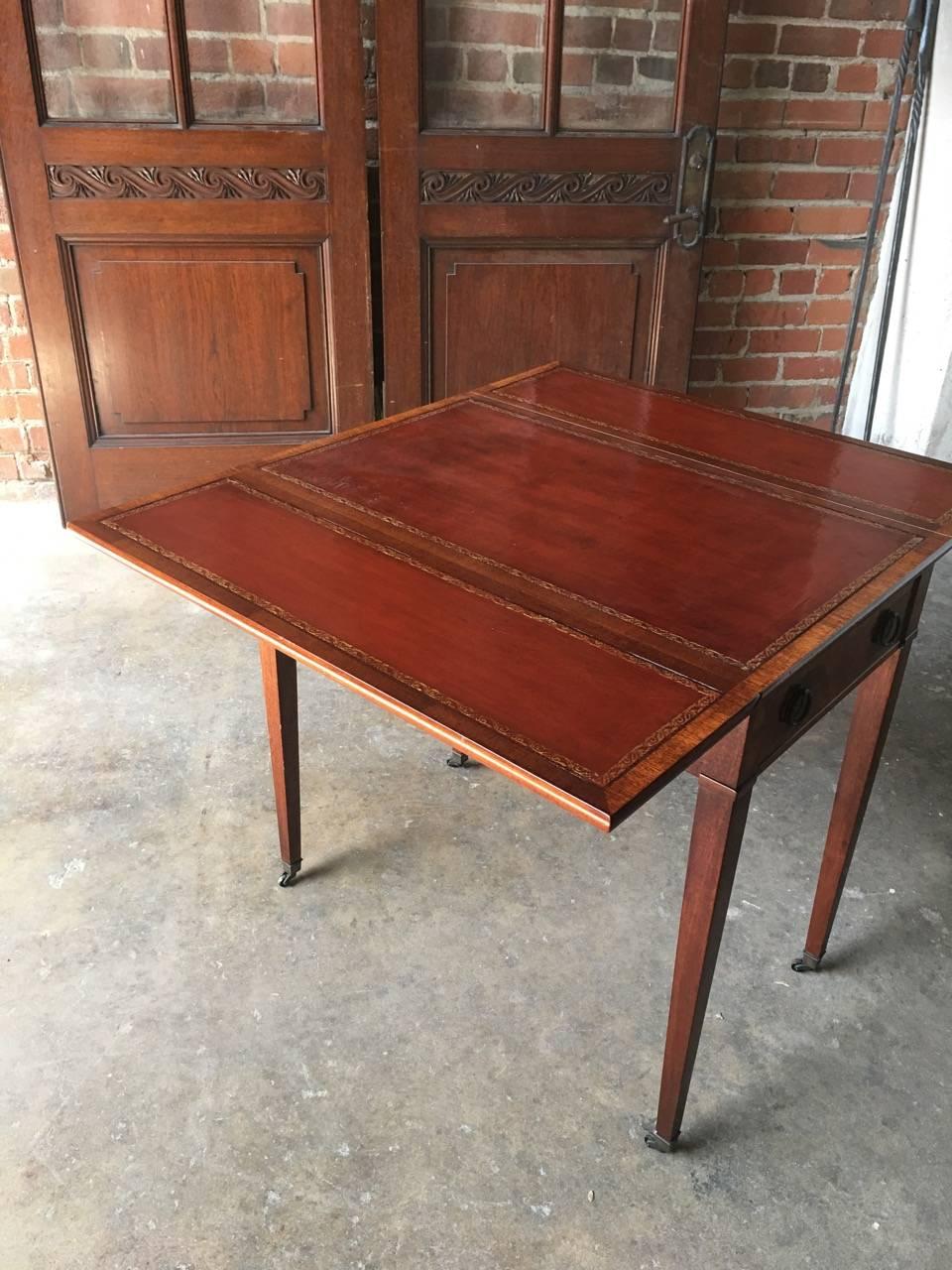 Elegant George III Pembroke table constructed of mahogany with inlay detail on the single drawer and opposing dummy false drawer. Features 3 cordovan leather surfaces with gold tooled leather borders on top and both drop leaf sides. Standing on