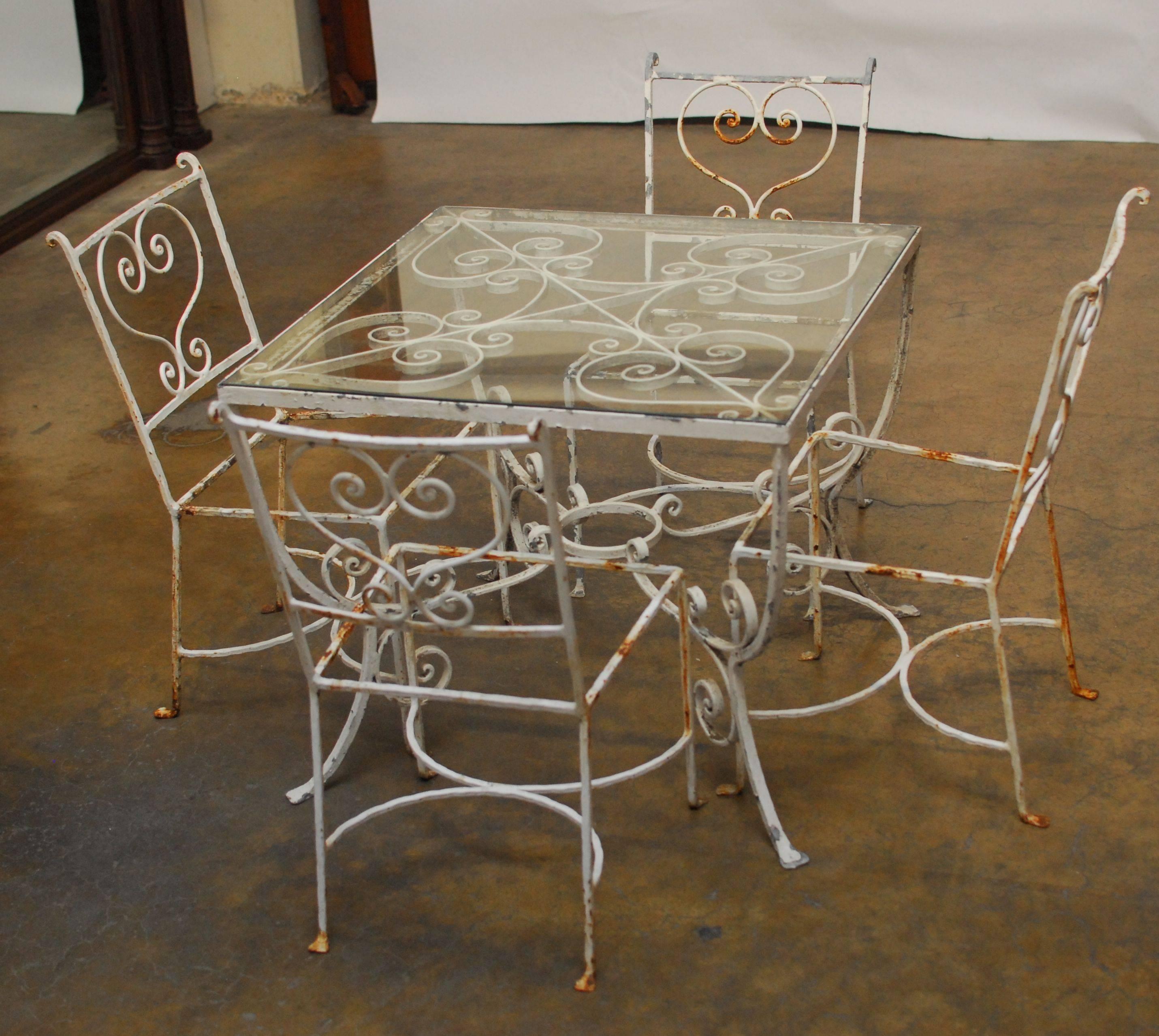 Lovely French garden patio set with 4 garden chairs and a glass top table featuring a plant shelf. The table and chairs have a French scrolled heart motif and sit on flat feet. Original distressed white chippy finish with minor surface rust. The