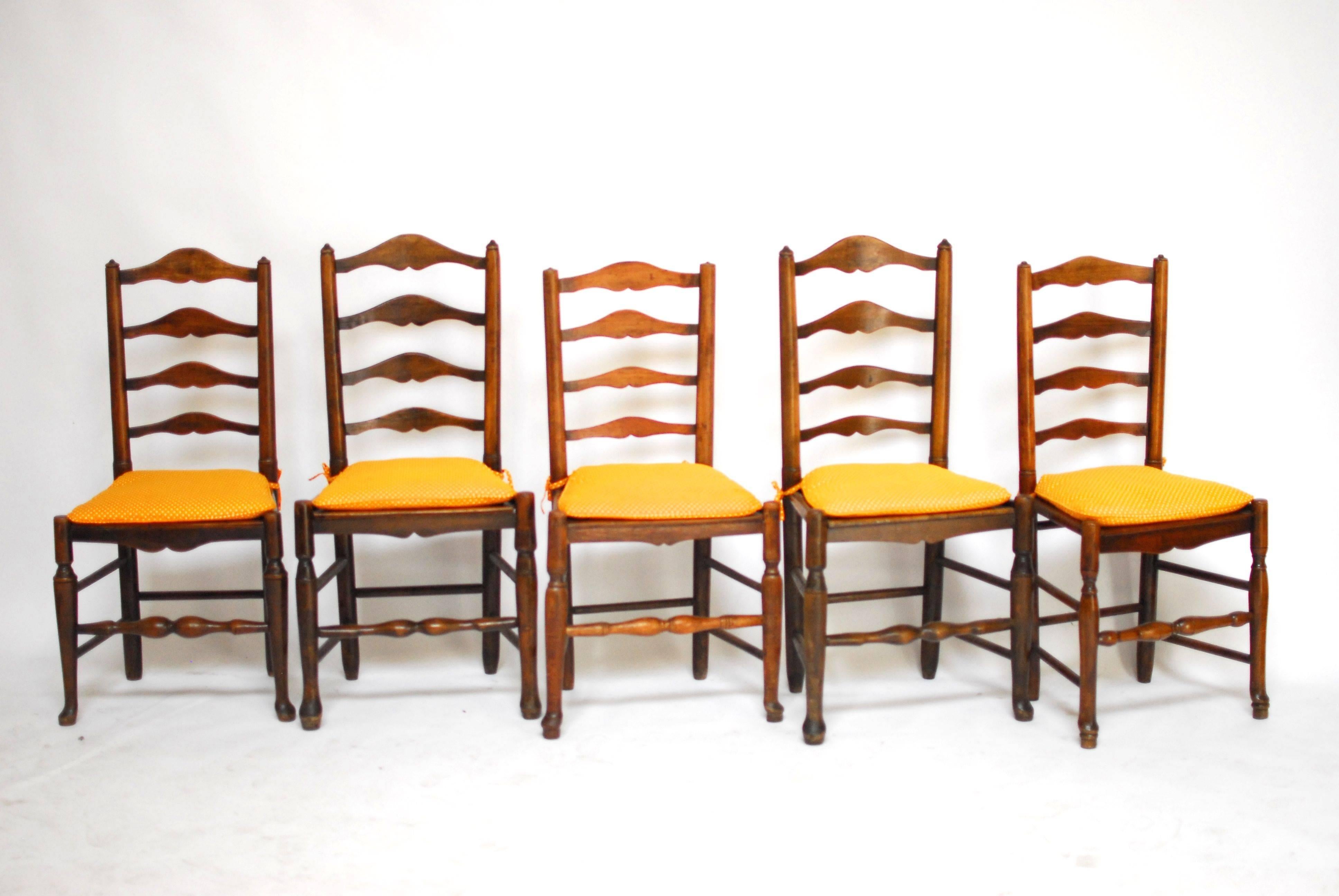 A set of five, rustic English country ladder back chairs from the 1850s made of elm and ash. Each chair has four shaped slats with tapered stiles and small finials on top. They have wood plank seats with turned legs and front stretchers sitting on