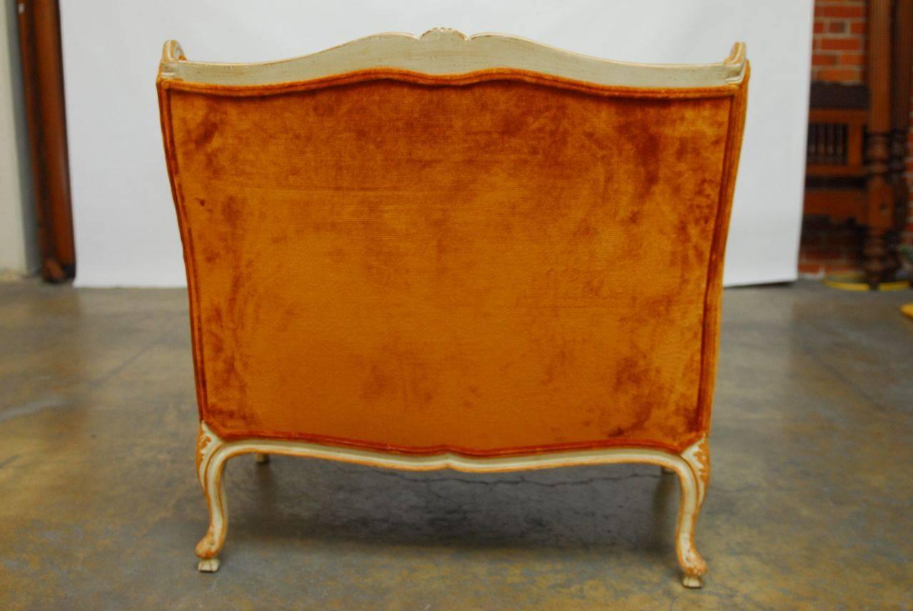 Fabulous French winged loveseat featuring plush burnt orange velvet upholstery. Hand-carved frame with cabriole legs and a serpentine crest finished in a cream lacquer with orange lacquer highlights. Double welt upholstery trim and thick down filled