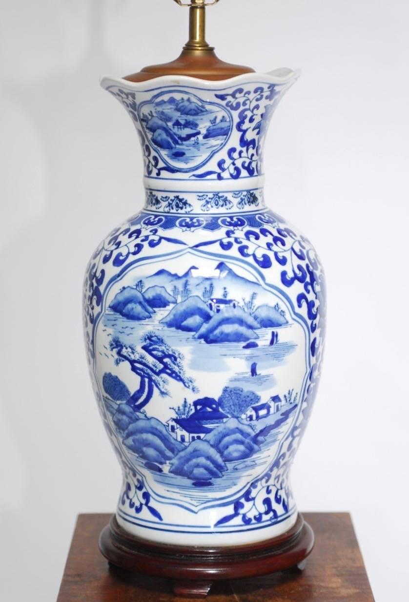 Large Chinese blue and white porcelain ginger jar table lamp featuring a baluster form jar with a scalloped lip. The body is decorated with an idyllic Asian scene on each side framed by floral and foliate decoration. Supported by a carved wooden