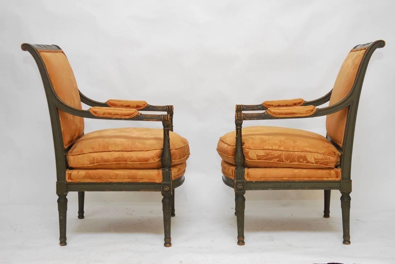 Rare pair of painted fauteuil armchairs made in the Directoire style featuring generous molded frames with a sweeping back. Carved in a neoclassical motif and supported by tapered column legs. Upholstered with a silk damask fabric in a rose gold