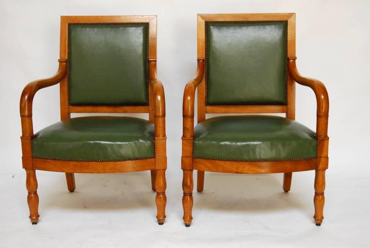 Impressive pair of French Empire Mahogany library chairs featuring square backs and gracefully curved arms that attach to a generous seat. Upholstered in a Napoleonic dark green with small brass nailhead trim. The chairs or fauteuils are supported