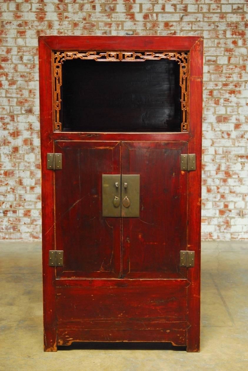 Impressive Chinese red lacquer cabinet with an open display shelf. Featuring an aged red lacquer finish and intricate carved fretwork decoration on the front panel. The case has two large doors that open to reveal a storage area with two drawers.