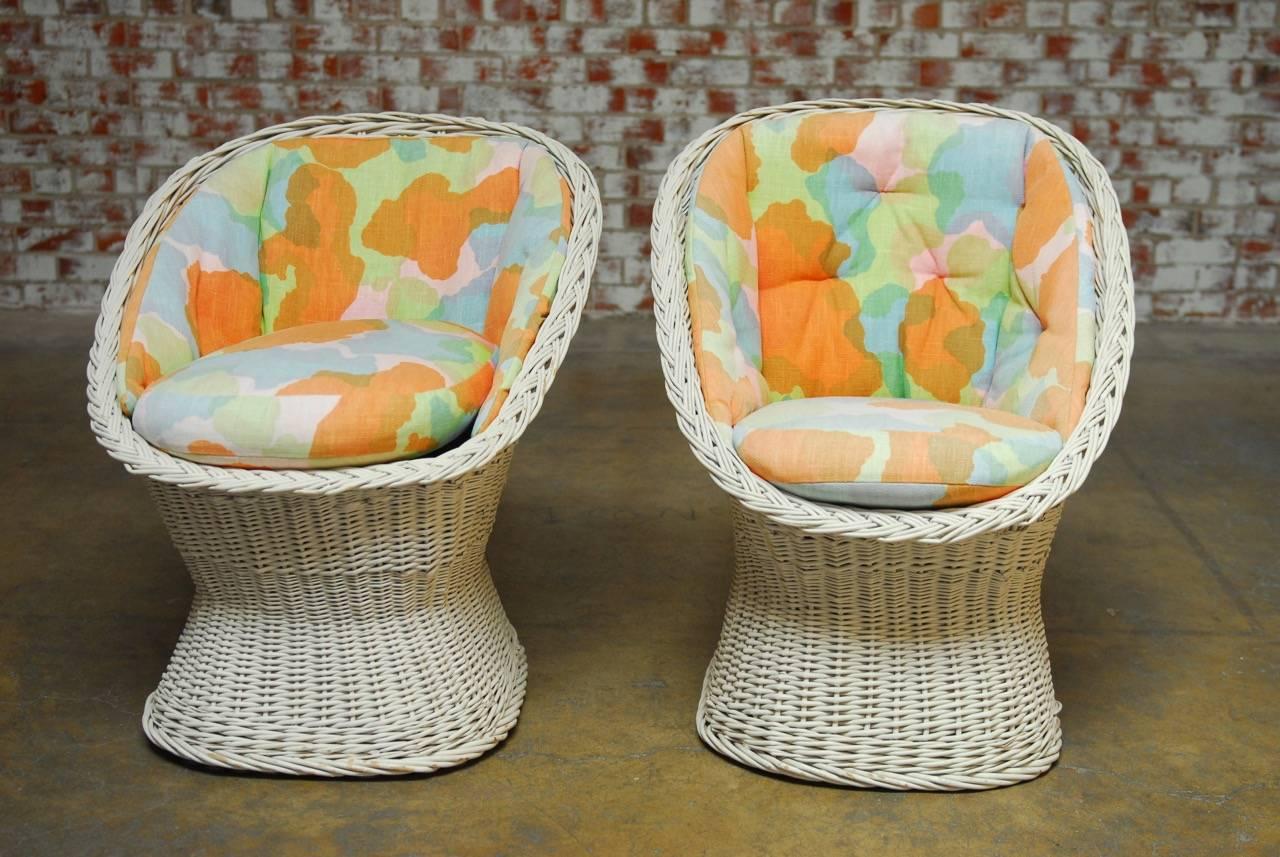 Charming pair of Mid-Century Modern French wicker egg cup chairs featuring a woven wicker frame with a flared bottom and a braided lip. Adorned with a multicolored linen fitted interior consisting of a wraparound back pad and a thick seat cushion.