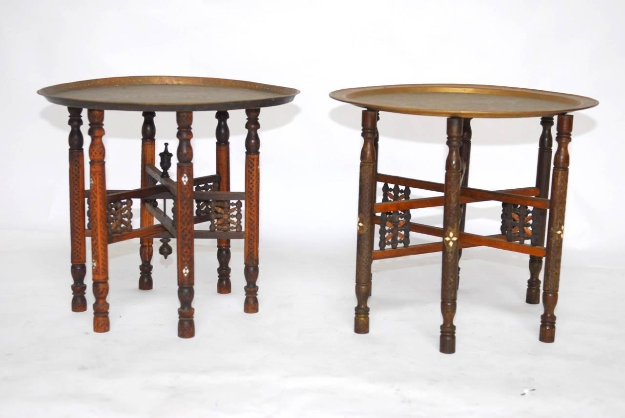 Traditional pair of Moroccan brass tray and inlaid wood folding drink tables. Featuring six-leg folding bases with intricate carved details and inlay. Topped with patinated etched brass tea trays. Each table base slightly varies in style and