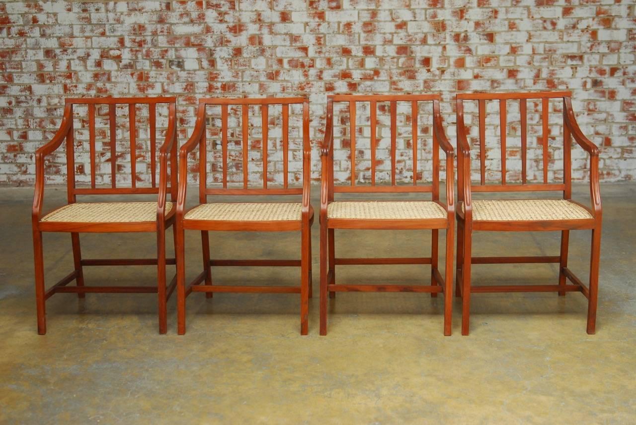 Stylish set of four slat back hand-carved dining chairs featuring a hand-caned seat. Each chair has long, graceful finger carved arms and is supported by elegant square legs with H shaped stretchers. Understated with a warm teak finish and excellent