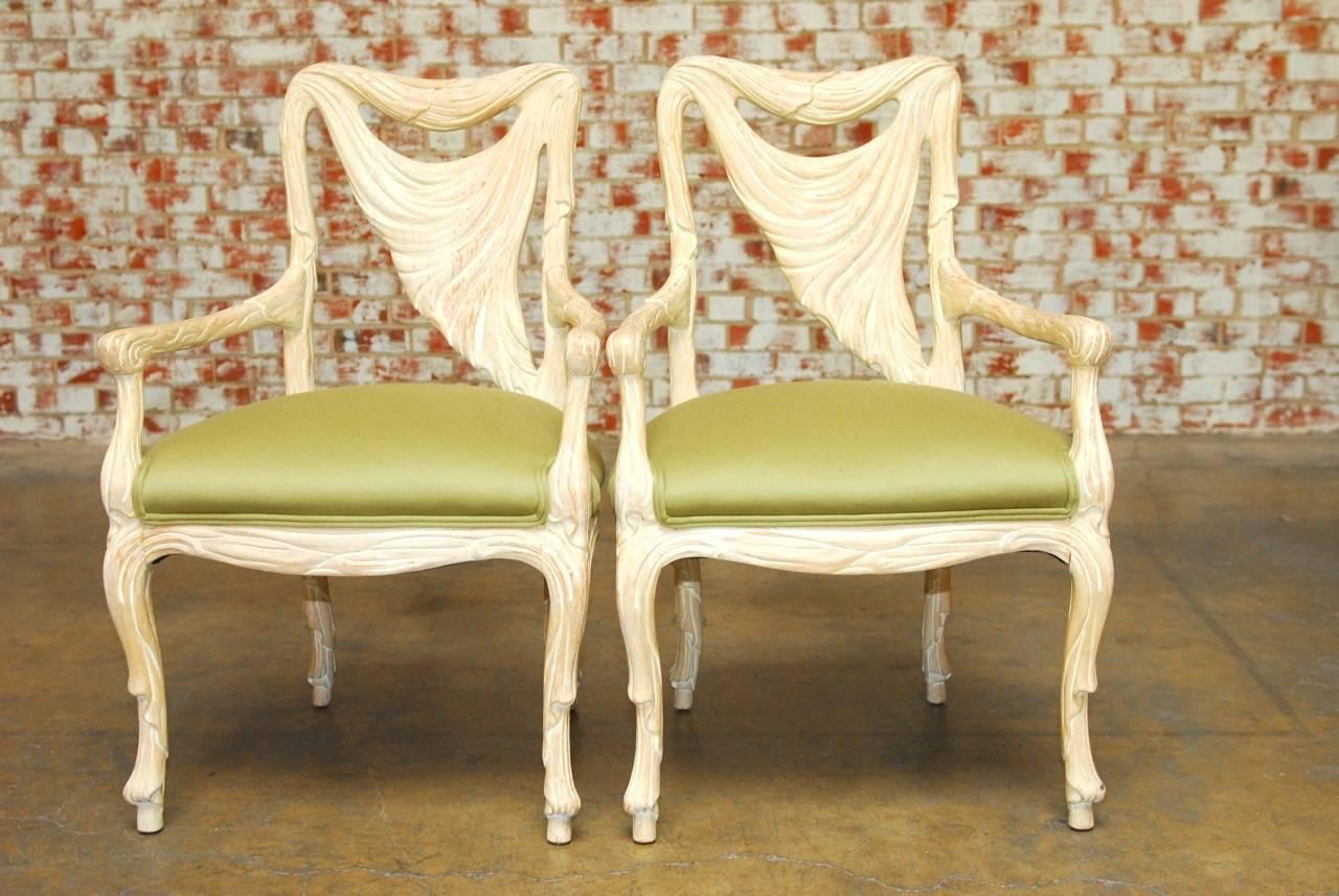 Stunning pair of Hollywood Regency armchairs featuring hand-carved frames with a faux-draped material motif. The arms, legs, and back are decorated with swag. The frames have been painted and have a beautifully distressed finish that showcases the