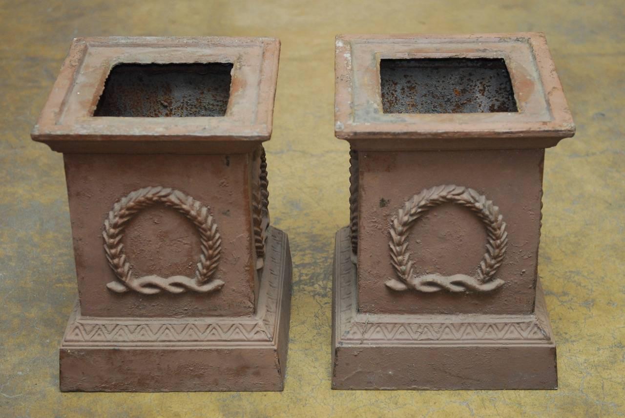 Handsome pair of cast iron square pedestals or garden urns made in the Neoclassical taste. Featuring an iron wreath decoration on all four sides with a patinated metal finish. These large square plinths could be used as planters or to display