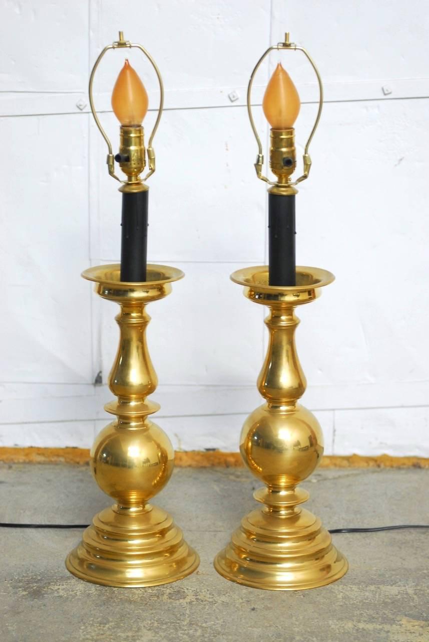 Stately pair of polished brass candlestick lamps having a turned baluster form. Each candlestick topped with a cupped bobeche and black faux candle. The sticks are heavy and thick with a rich luster. Surmounted by black fabric shades with a gold