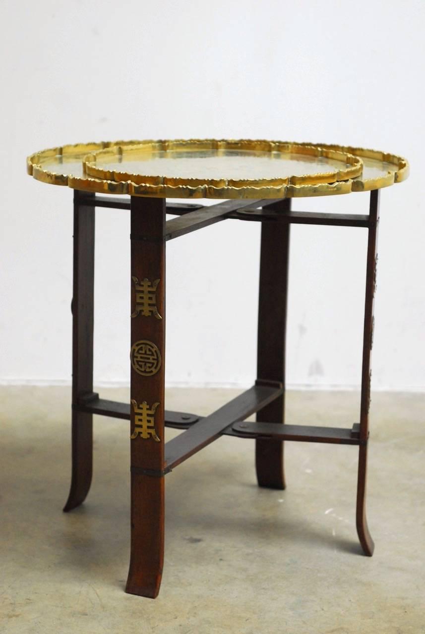 Interesting Asian folding brass tea tray table or drink table. Featuring a folding four leg base supporting two round trays. Each brass tray is incised with a decorative scene and a galleried edge with a scalloped detail. The smaller tray measures