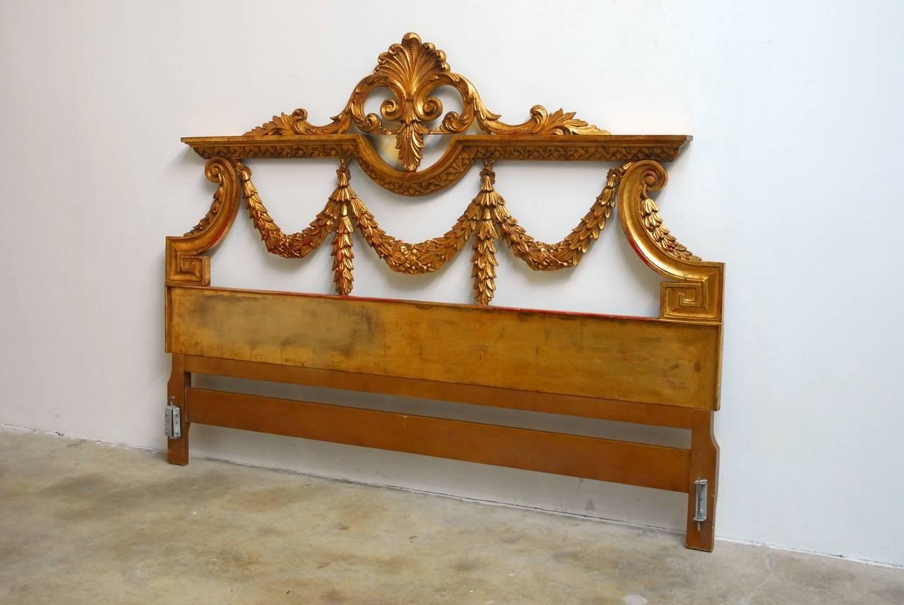 Majestic Italian neoclassical style carved headboard featuring a hand-applied gold leaf gilt finish over a deep red base. Made in the neoclassical taste showcasing carved swag laurel wreaths bordered by greek key motif scrolls. Surmounted by a thick