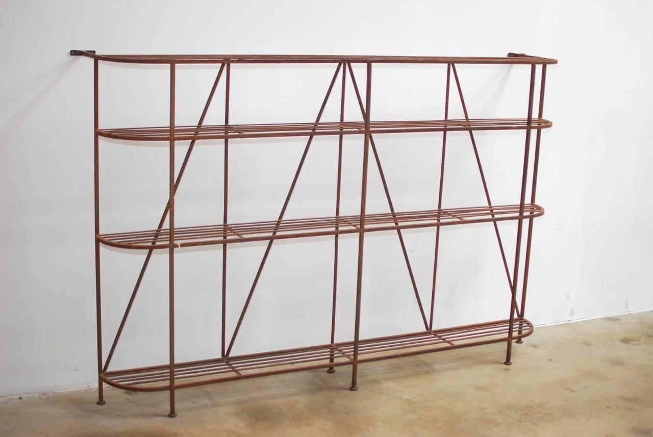 Industrial French Art Deco iron display shelf or bakers rack. Features four large shelves with curved ends constructed from solid square iron frame with a patinated metal finish. Each side has wall anchors on the top and the shelf is supported by