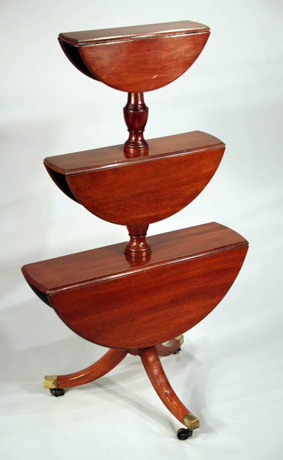 Small-scale George III Sheraton three-tier dumbwaiter in mahogany, having 
circular shelves with drop leaves, vasiform pillars and down swept legs 
ending in brass casters.
English, circa 1790.
 