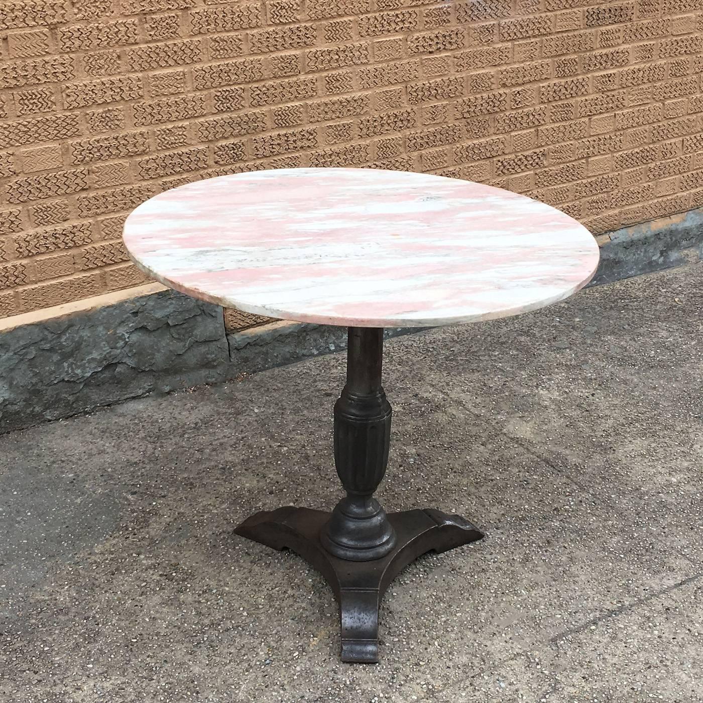 Bistro / café / dining table has a vintage, round marble top with pink and gray hues atop a decorative, cast iron pedestal base in a raw steel finish, circa 1930s.