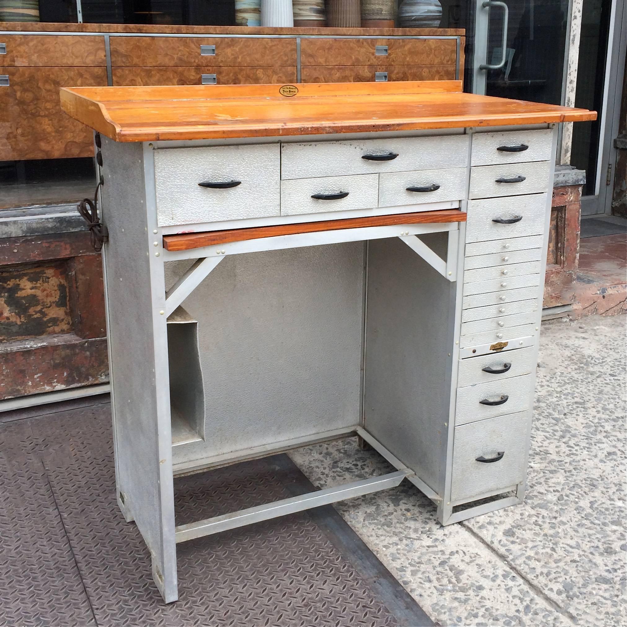 Industrial, watchmaker’s desk features a stamped aluminum base with 18 drawers of various size and a maple top. There is also a maple framed canvas ledge that pulls out below the center drawers to catch small parts and 2 electrical outlets on the