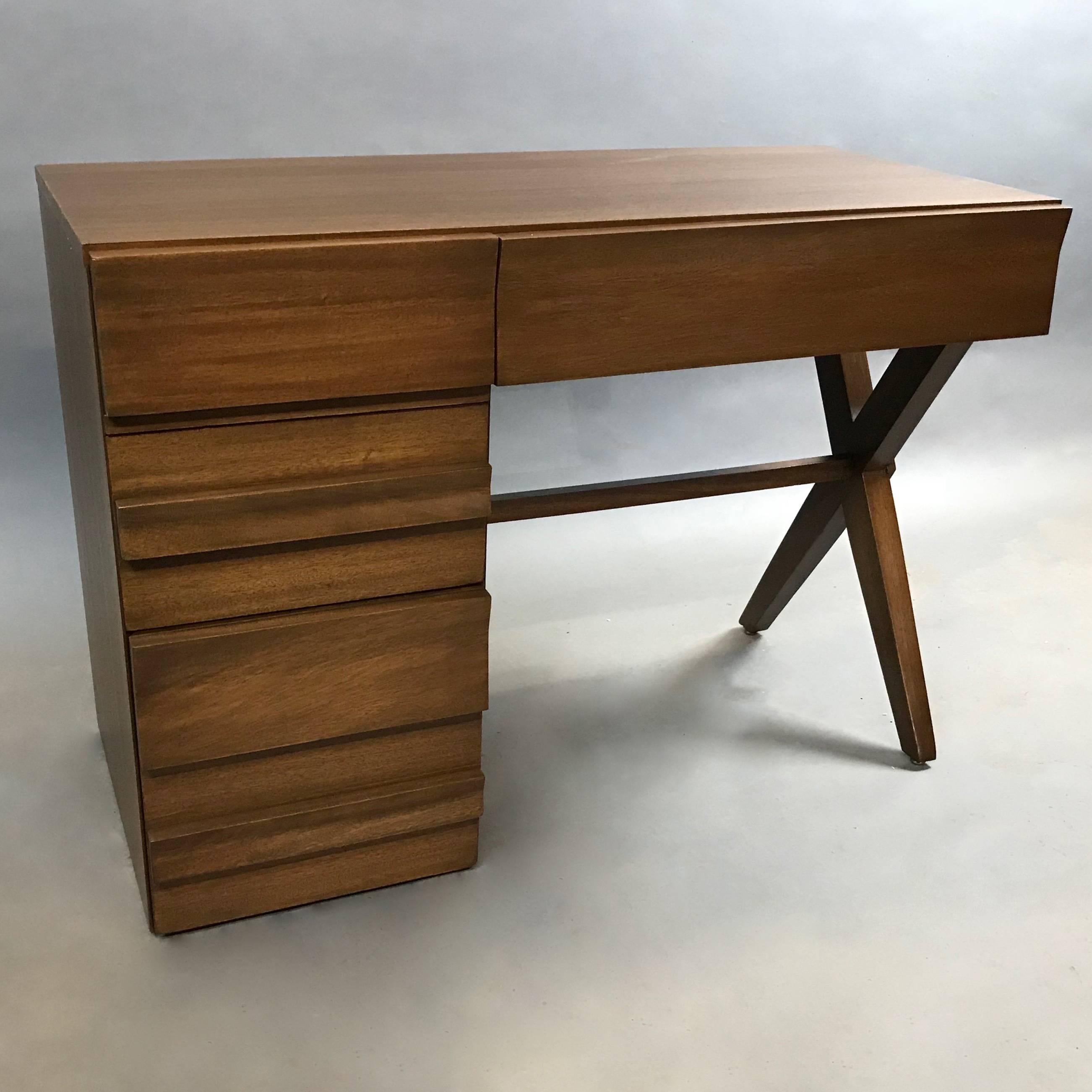 Highly styled, compact, Mid-Century Modern, mahogany pedestal desk features three decoratively hidden drawers with a carved undulating pattern throughout, detailed X-base and pull-out shelf. Seat clearance is 24.5in ht. The bottom drawer is deep