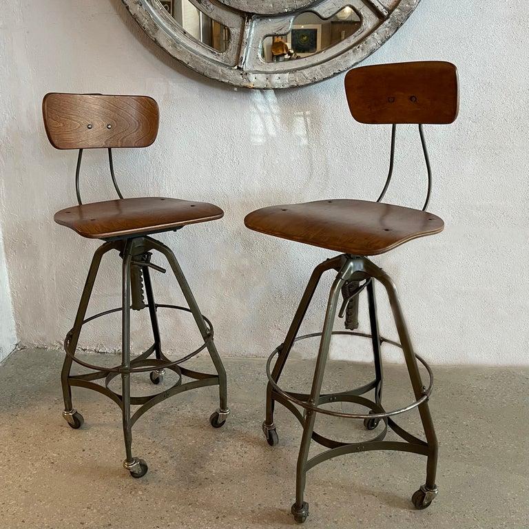 Pair of 1940's industrial drafting stools by Toledo Metal Furniture Co. feature height adjustable, gray green painted steel, A frame bases with bent maple ply, swivel seats and backs. The stools are height adjustable from 28 - 33 inches and roll