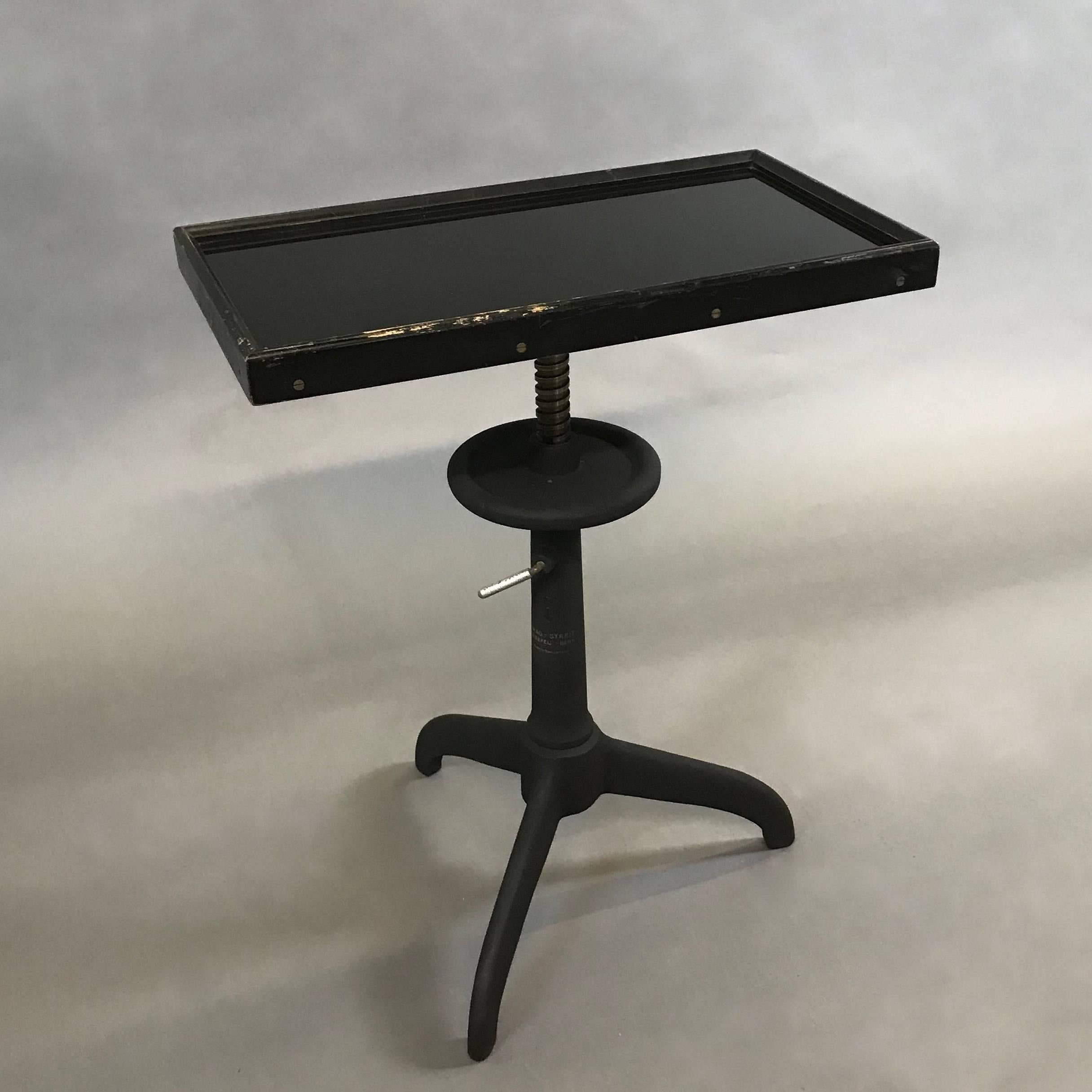 Industrial, optometry examination table by Bausch & Lomb features a black glass top in a wood frame atop a height-adjustable, pedestal cast iron and steel base.