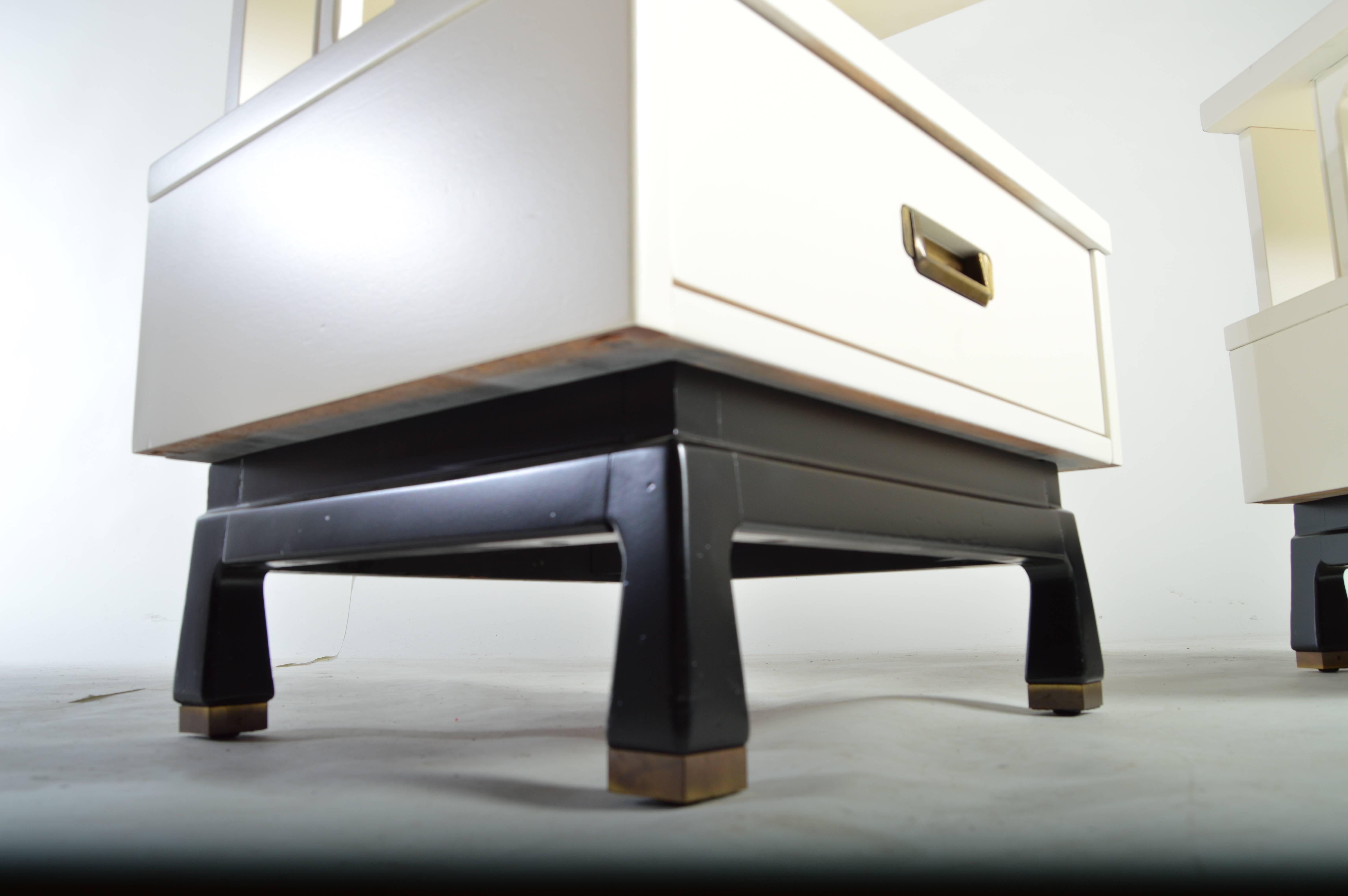 Fabulous pair of Mid-Century nightstands or end tables having brass pulls and feet by American of Martinsville. Freshly refinished in bone lacquer with an  ebonized base.  