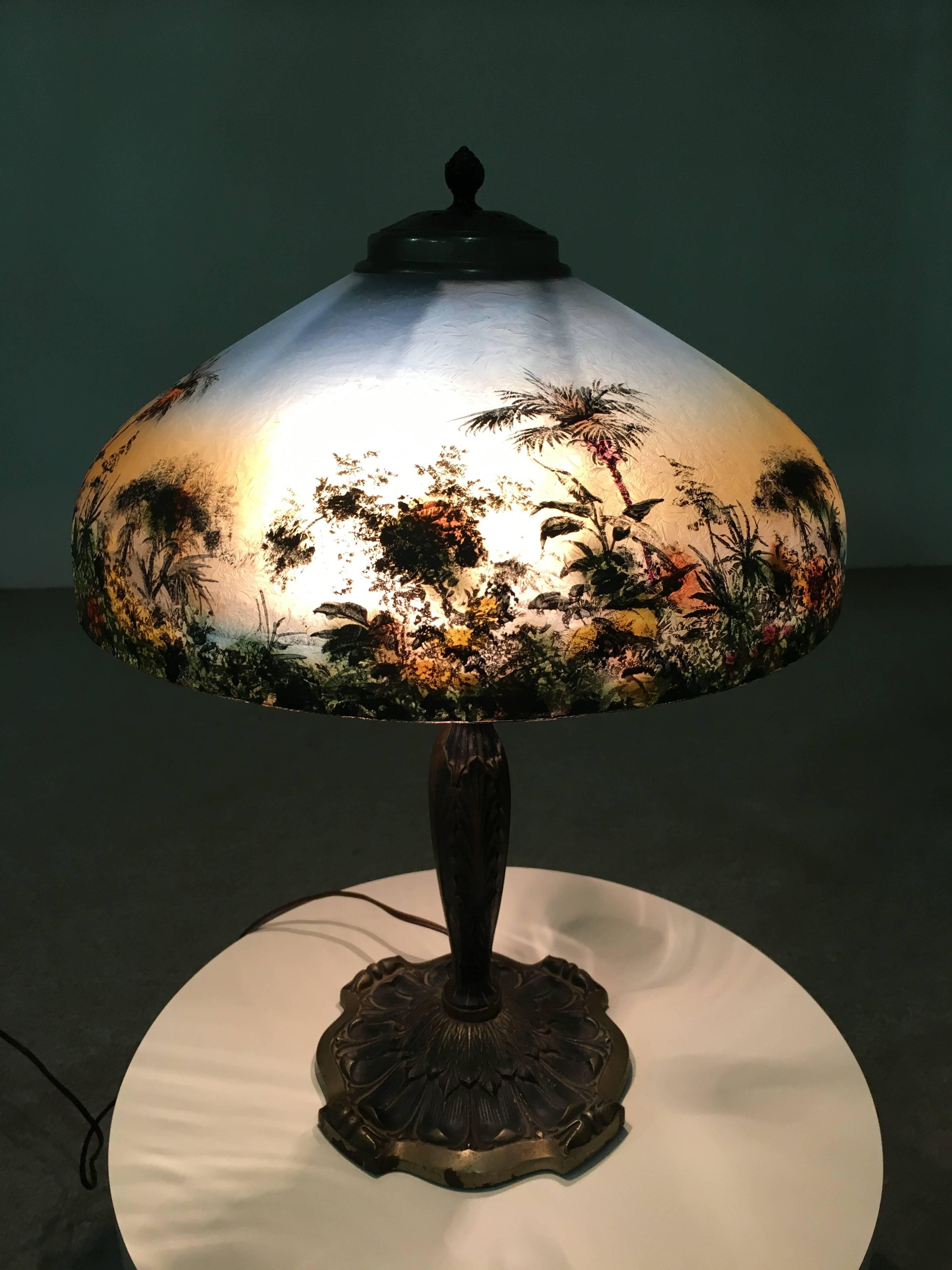 A beautiful 1930s Pittsburgh reverse painted table lamp having lush tree tropical foliage with palm trees and birds throughout atop a bronzed base. Partial Pittsburgh label still visible underneath the shade. Very good condition having no cracks or